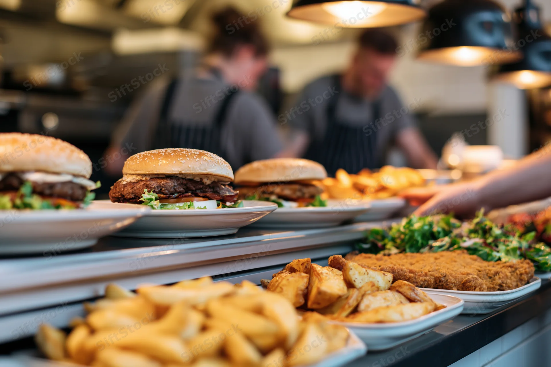 Plated burgers and fries on a restaurant pass counter with chefs in the background