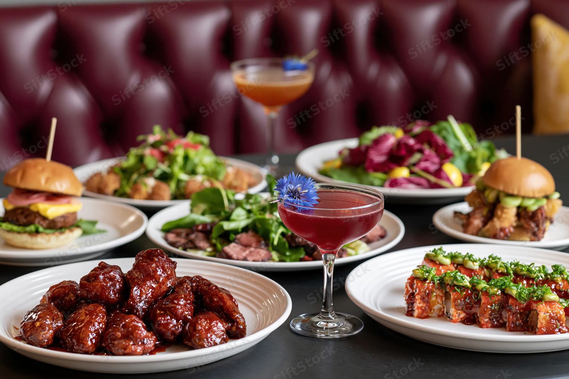 Restaurant table spread with multiple dishes including glazed meatballs, burgers, salads, sushi, and cocktails on dark surface with tufted leather booth in background