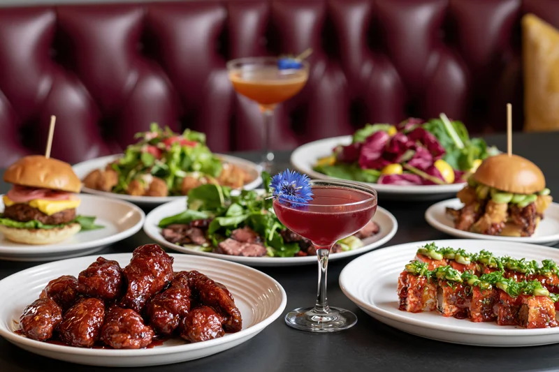 Restaurant table spread with multiple dishes including glazed meatballs, burgers, salads, sushi, and cocktails on dark surface with tufted leather booth in background