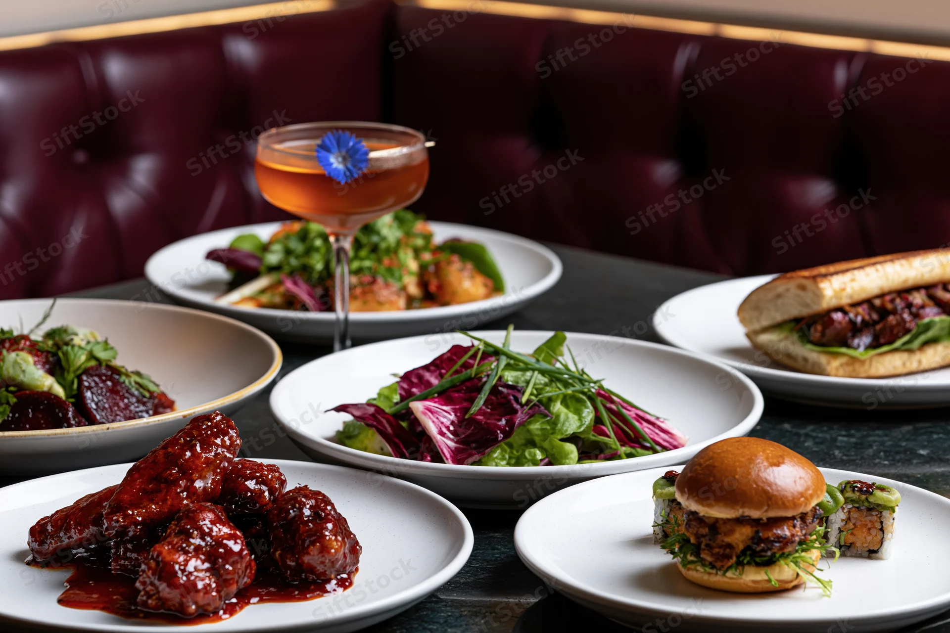 Multiple plated dishes including glazed wings, salad, burger, and cocktail on a restaurant table with tufted red leather booth in background