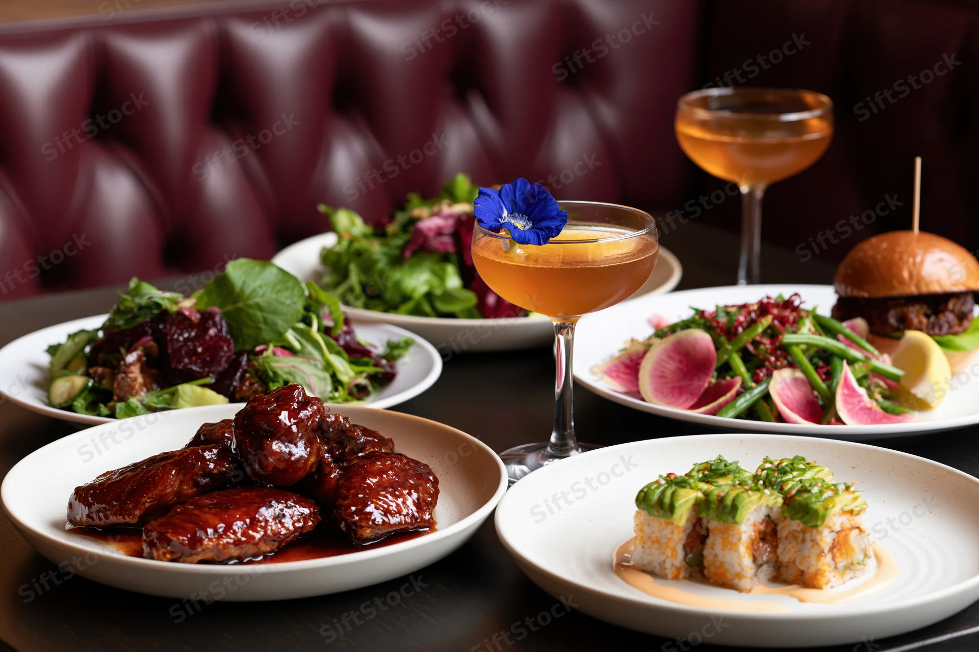 Restaurant table with multiple plated dishes including glazed wings, sushi, salad, burger and cocktails arranged for dinner service