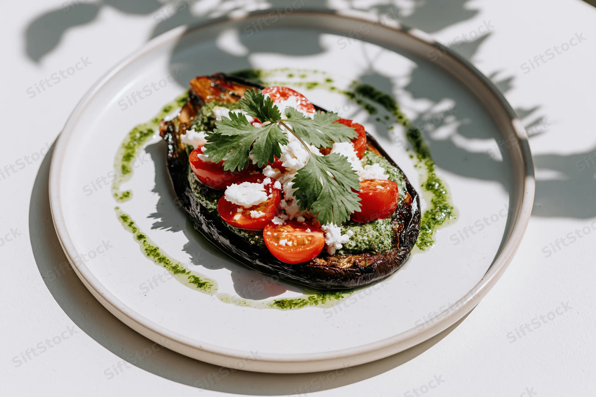 Roasted eggplant topped with tomatoes, feta, cilantro and pesto sauce on a white ceramic plate under dappled sunlight