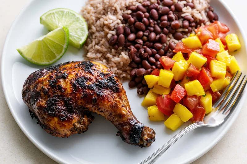Roasted jerk chicken leg quarter on a white plate with rice, black beans, mango salsa and lime wedges