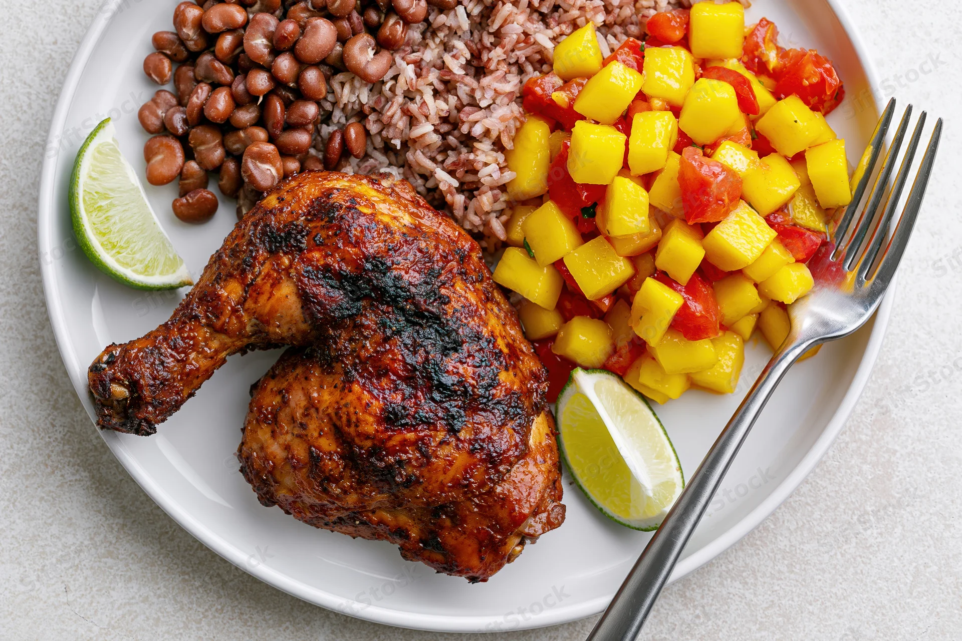 Roasted jerk chicken leg quarter on a white ceramic plate with beans, rice, mango salsa, lime wedges, and a fork