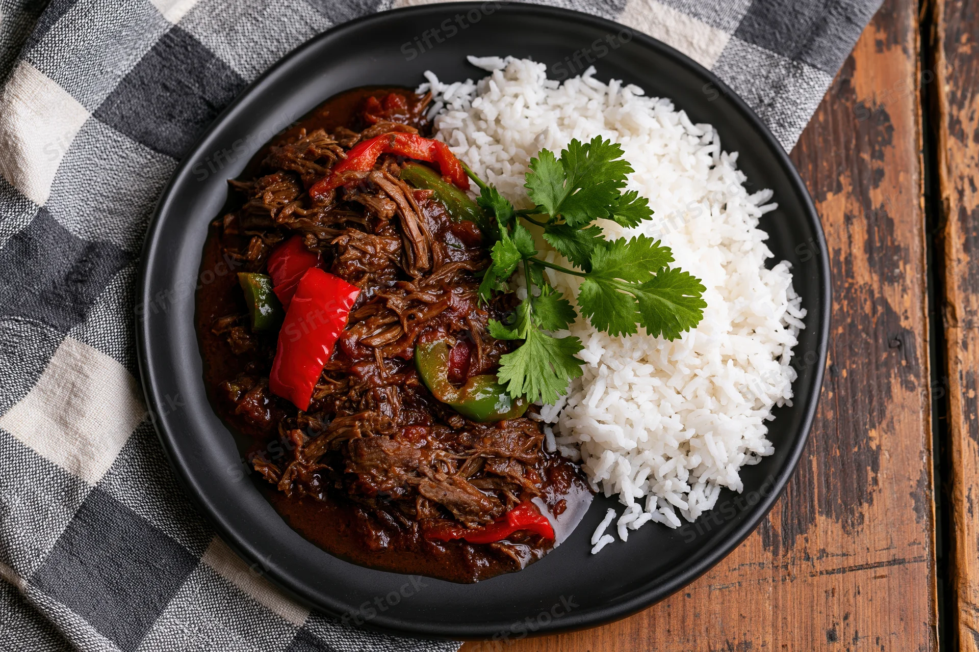 Shredded beef stew with rice and cilantro on a black plate over a checkered napkin and wooden table
