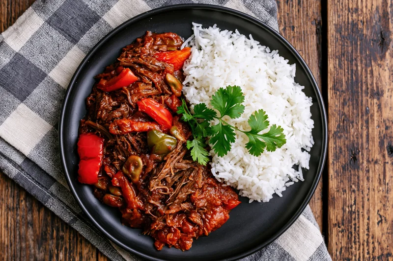 Shredded beef stew with rice and bell peppers on a black plate over a checkered napkin on a wooden table
