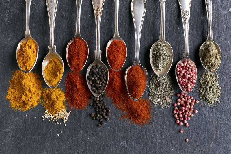 Row of antique silver spoons filled with assorted spices arranged on dark slate surface