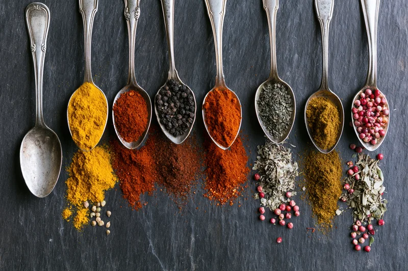 Row of antique silver spoons filled with assorted spices arranged on dark slate surface