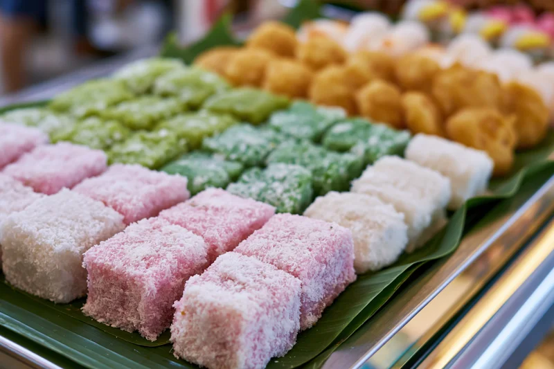 Rows of colorful southeast asian kueh sweets arranged on banana leaf tray at market display