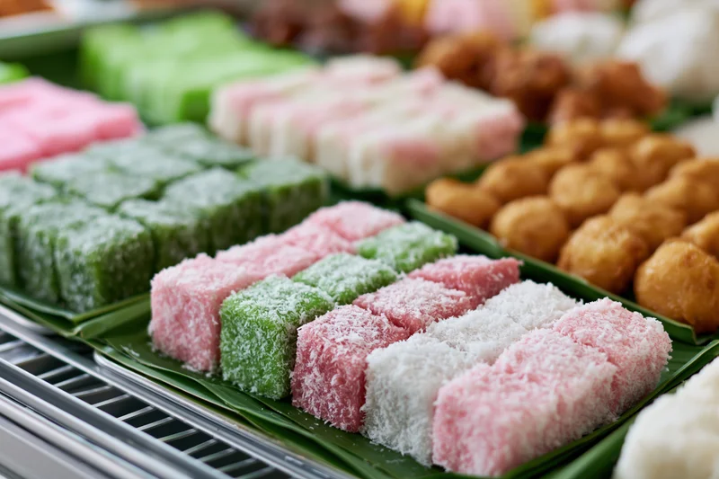 Rows of colorful southeast asian kueh sweets arranged on banana leaves in a market display