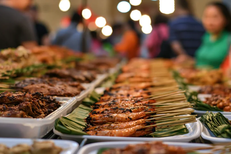 Rows of grilled prawn and meat skewers arranged on white trays lined with banana leaves at a bustling outdoor market stall