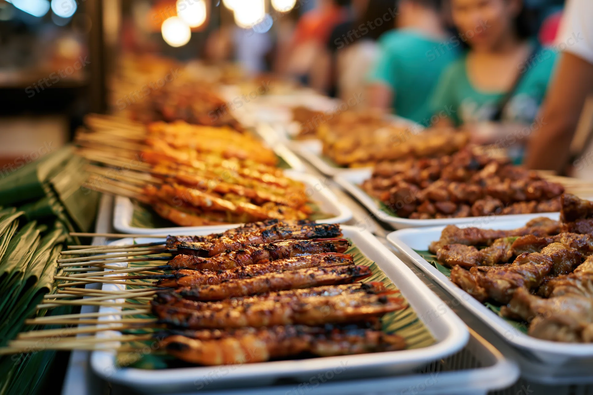 Rows of grilled meat and prawn skewers on banana leaves in white trays at a night market stall