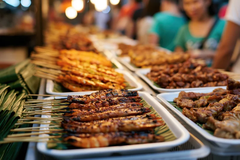 Rows of grilled meat and prawn skewers on banana leaves in white trays at a night market stall
