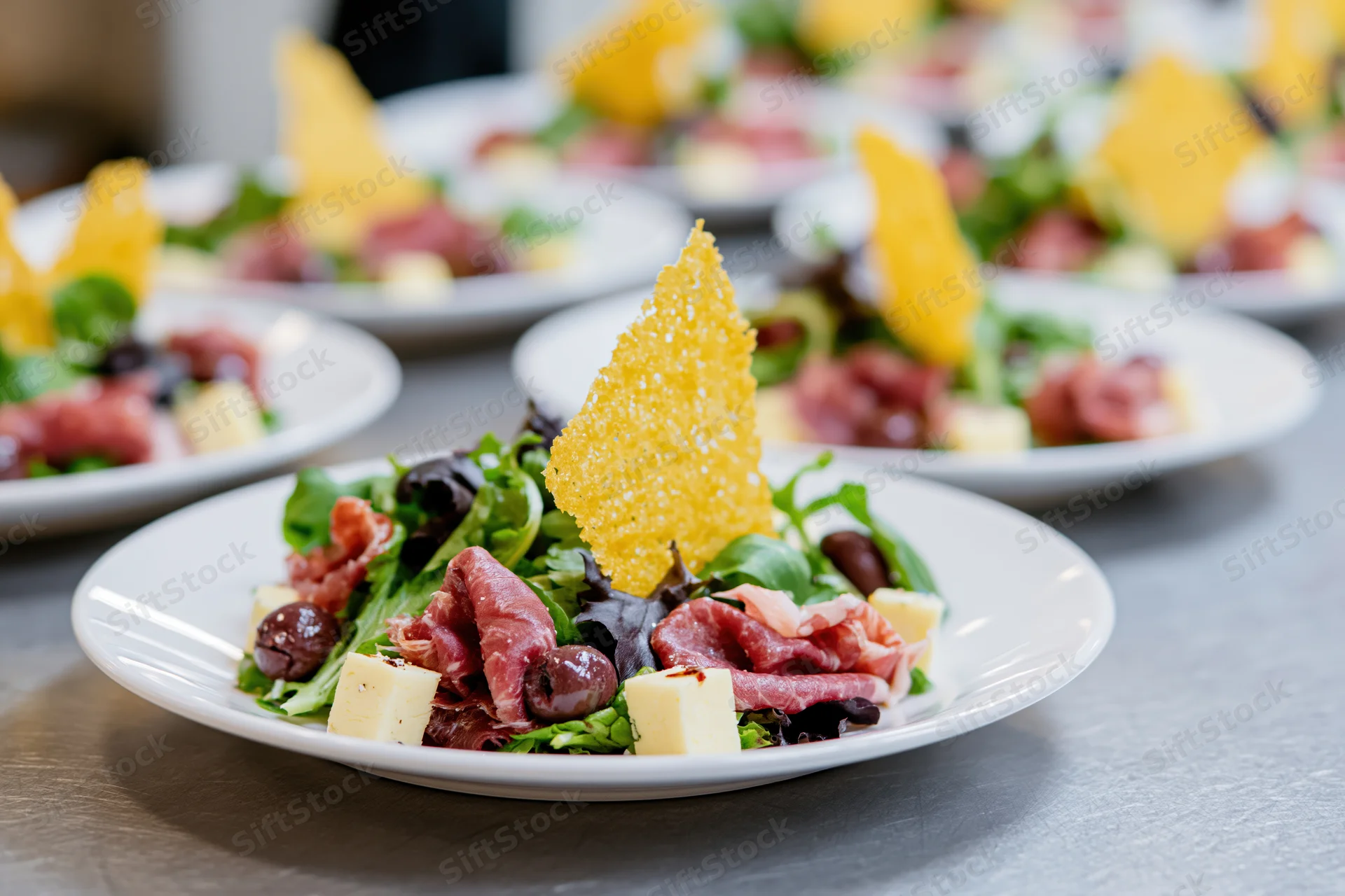 Rows of identical appetizer salads with prosciutto, cheese, olives and crispy garnish on white plates arranged on a stainless steel table