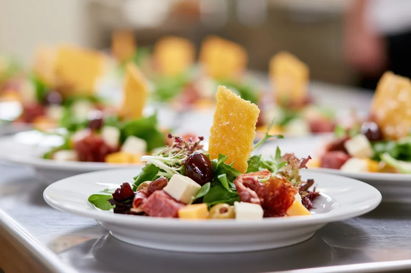Rows of identical appetizer salads with cheese, meat, and crackers on white plates arranged on a stainless steel tray