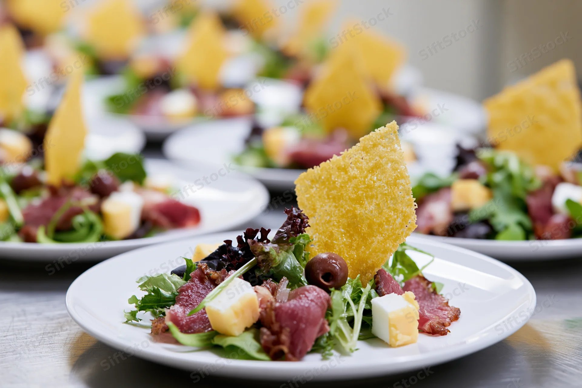 Rows of identical appetizer salads with cheese, olives, and crispy garnish on white plates arranged on a stainless steel table