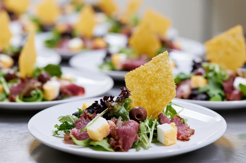 Rows of identical appetizer salads with cheese, olives, and crispy garnish on white plates arranged on a stainless steel table