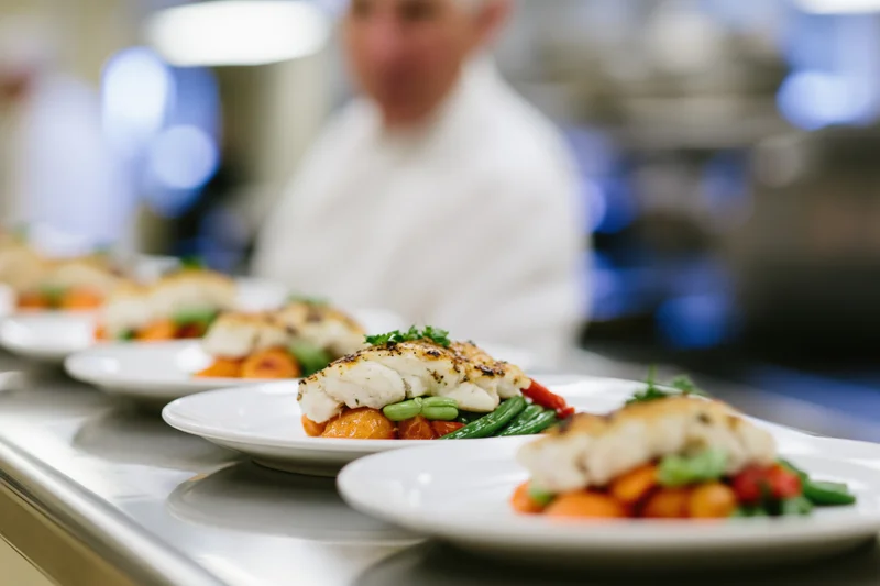 Rows of identical plated fish dishes lined up on a stainless steel surface in a restaurant kitchen with a chef in the background
