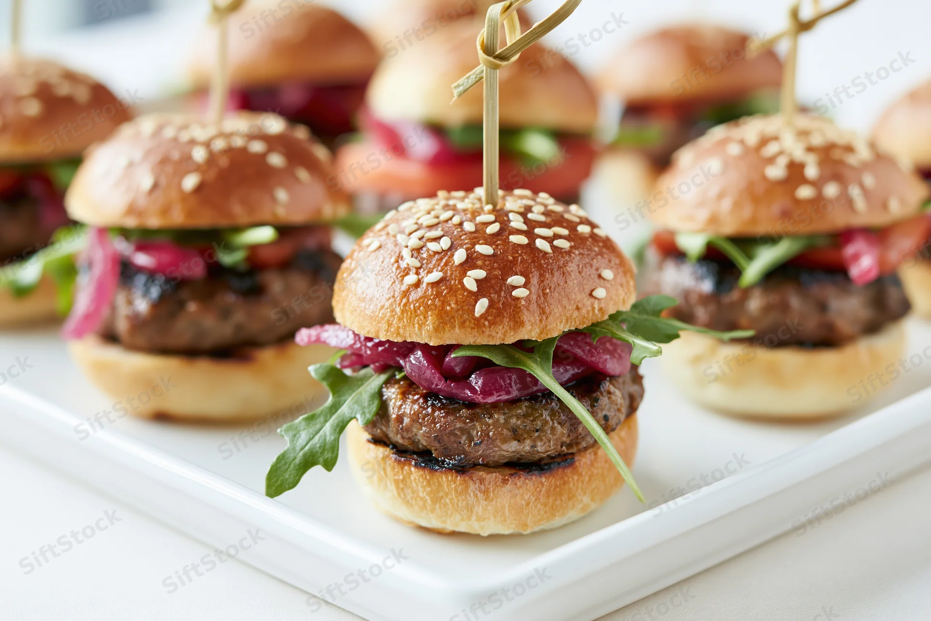 Mini beef sliders with sesame buns and arugula on a white rectangular plate
