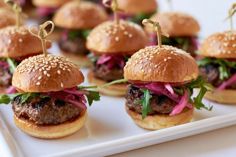 Mini beef sliders with sesame buns, pickled red onions and arugula on a white platter