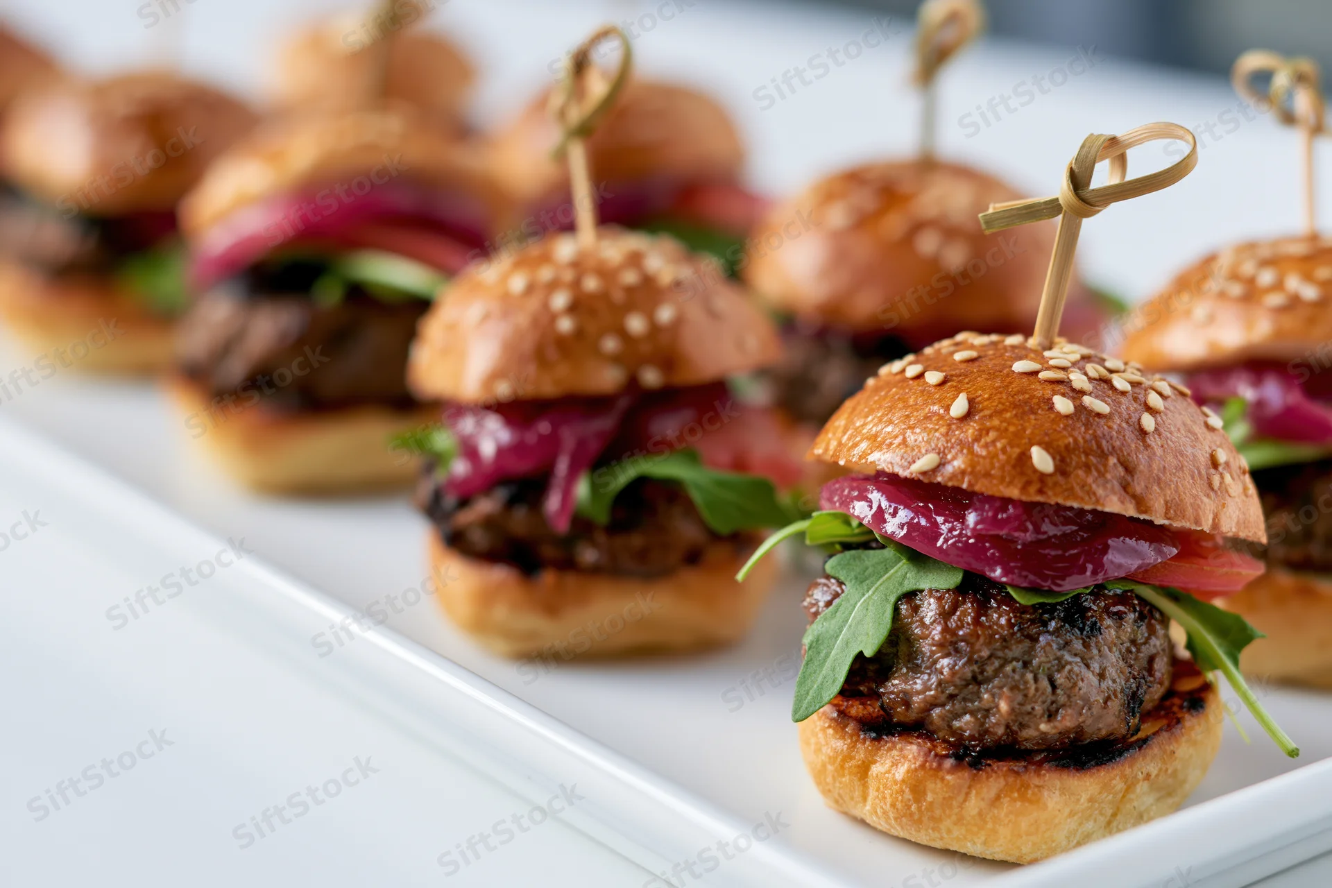 Mini beef sliders with sesame buns and arugula arranged in rows on a white rectangular platter