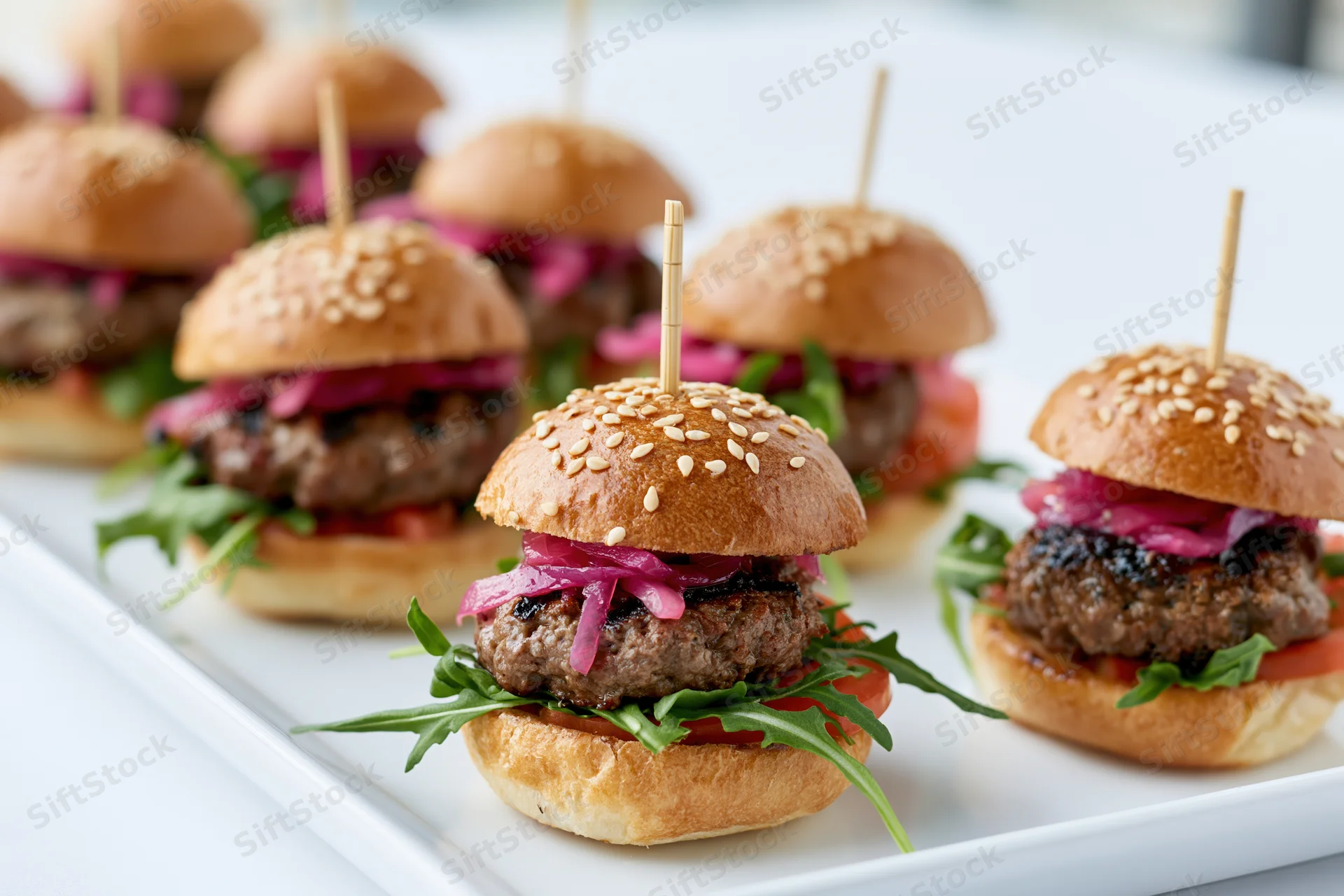 Mini beef sliders with pickled red onions and arugula on a white rectangular platter