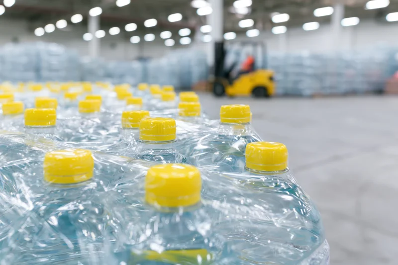 Shrink-wrapped plastic water bottles with yellow caps arranged in rows inside a warehouse with a forklift in the background