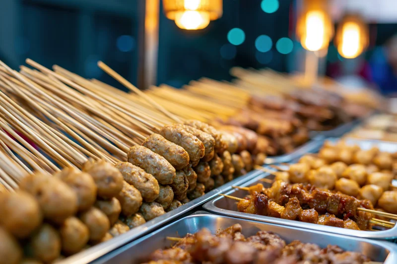 Rows of skewered meatballs and satay displayed in stainless steel trays at a night market