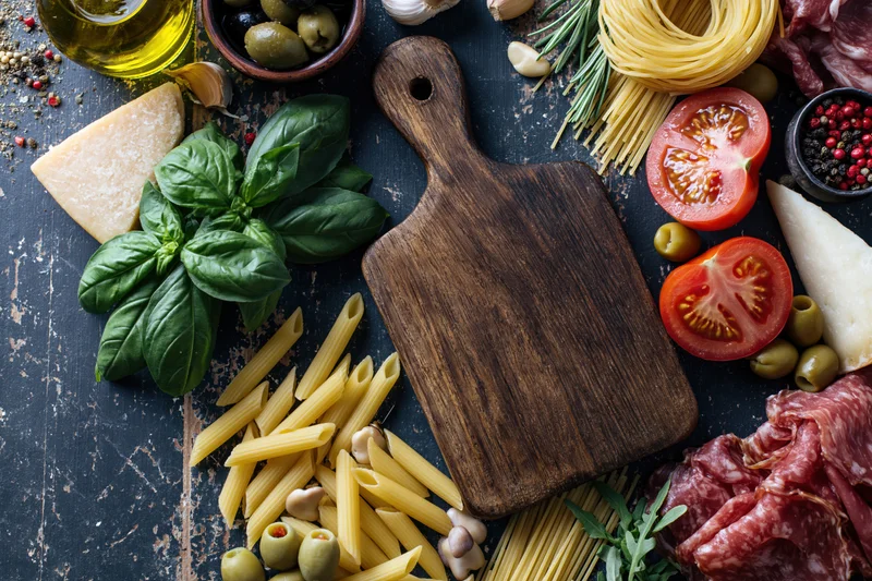 Rustic wooden cutting board surrounded by fresh italian ingredients on dark textured surface