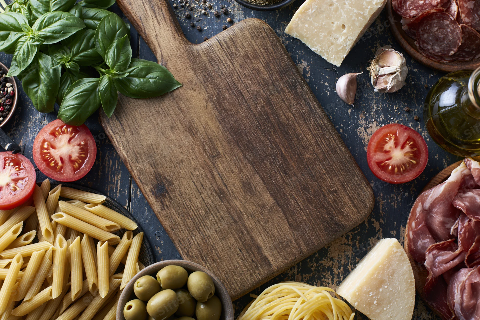 Wooden cutting board surrounded by pasta, cheese, tomatoes, basil, olives, garlic, salami and prosciutto on dark textured surface