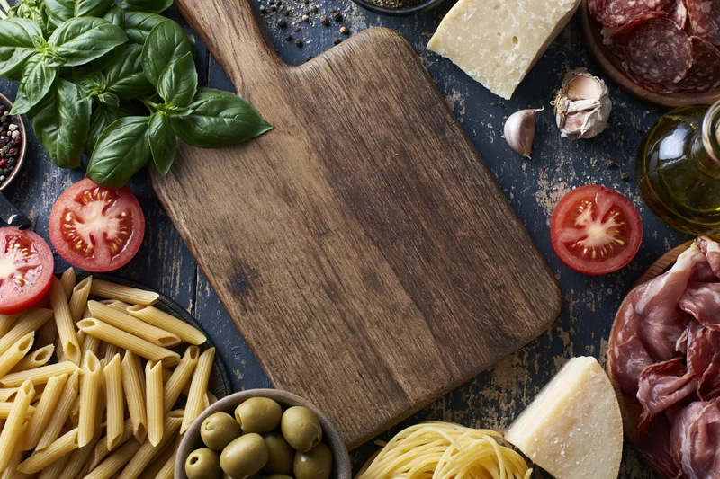 Wooden cutting board surrounded by pasta, cheese, tomatoes, basil, olives, garlic, salami and prosciutto on dark textured surface