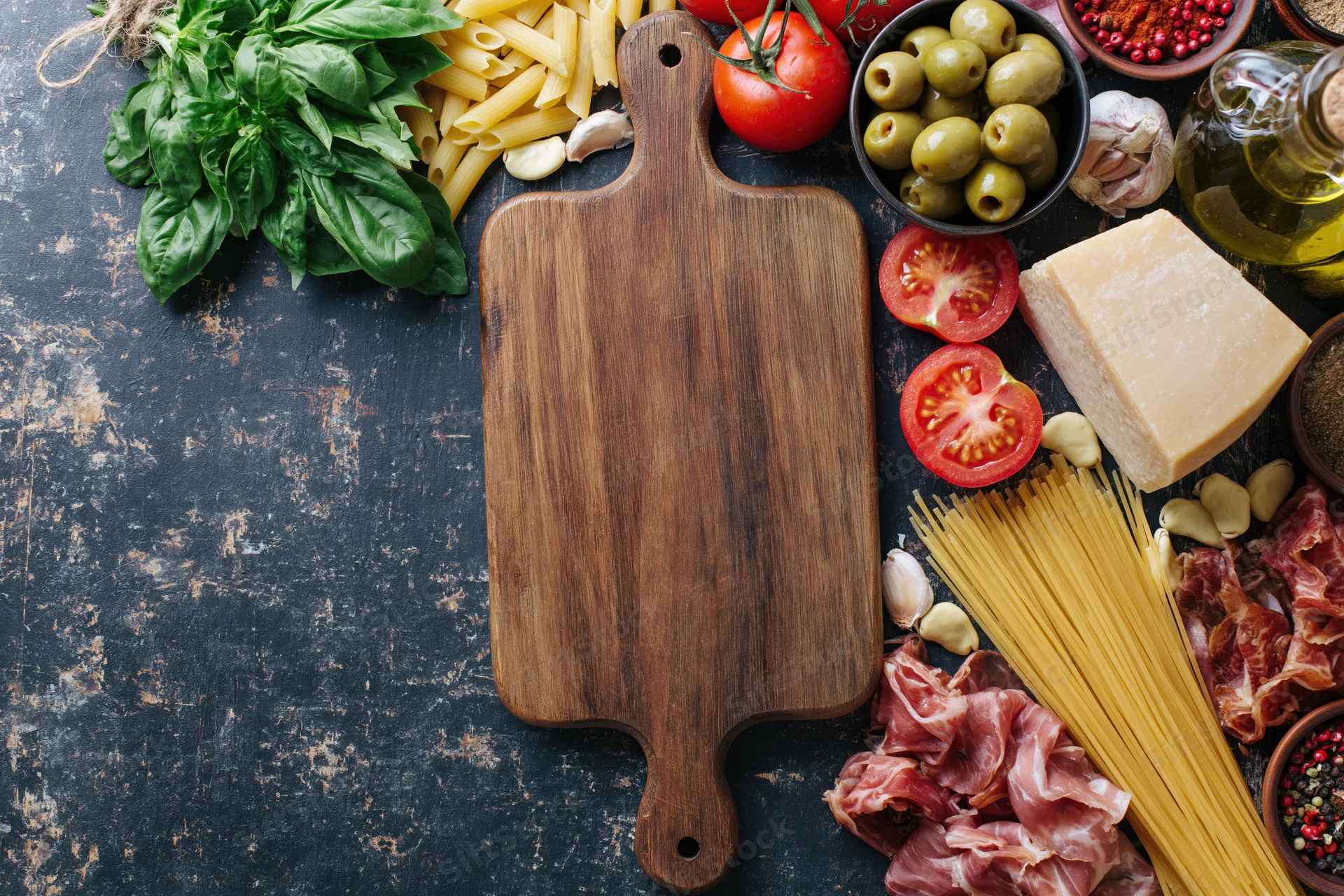 Wooden cutting board surrounded by fresh italian ingredients on dark textured surface