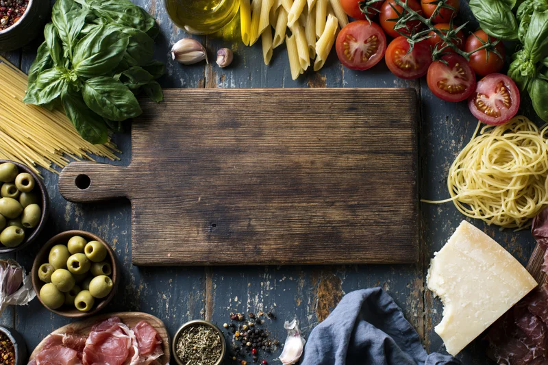 Rustic wooden cutting board centered on dark weathered table surrounded by fresh italian ingredients for cooking
