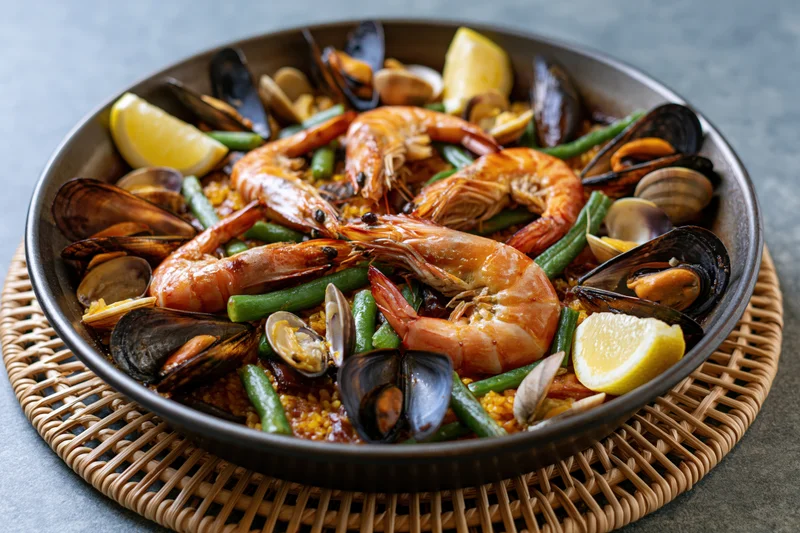 Seafood paella with shrimp, mussels, clams, green beans and lemon in a dark wide steel pan on a woven mat