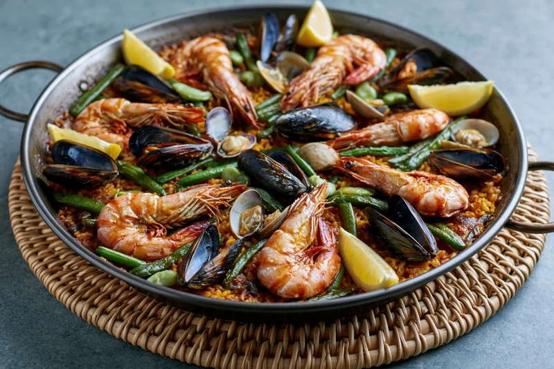 Seafood paella with shrimp, mussels, clams, green beans and lemon wedges in a wide dark steel pan on a woven mat