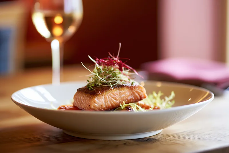 Seared salmon fillet topped with microgreens and red garnish on a white plate on a wooden table with a wine glass in the background