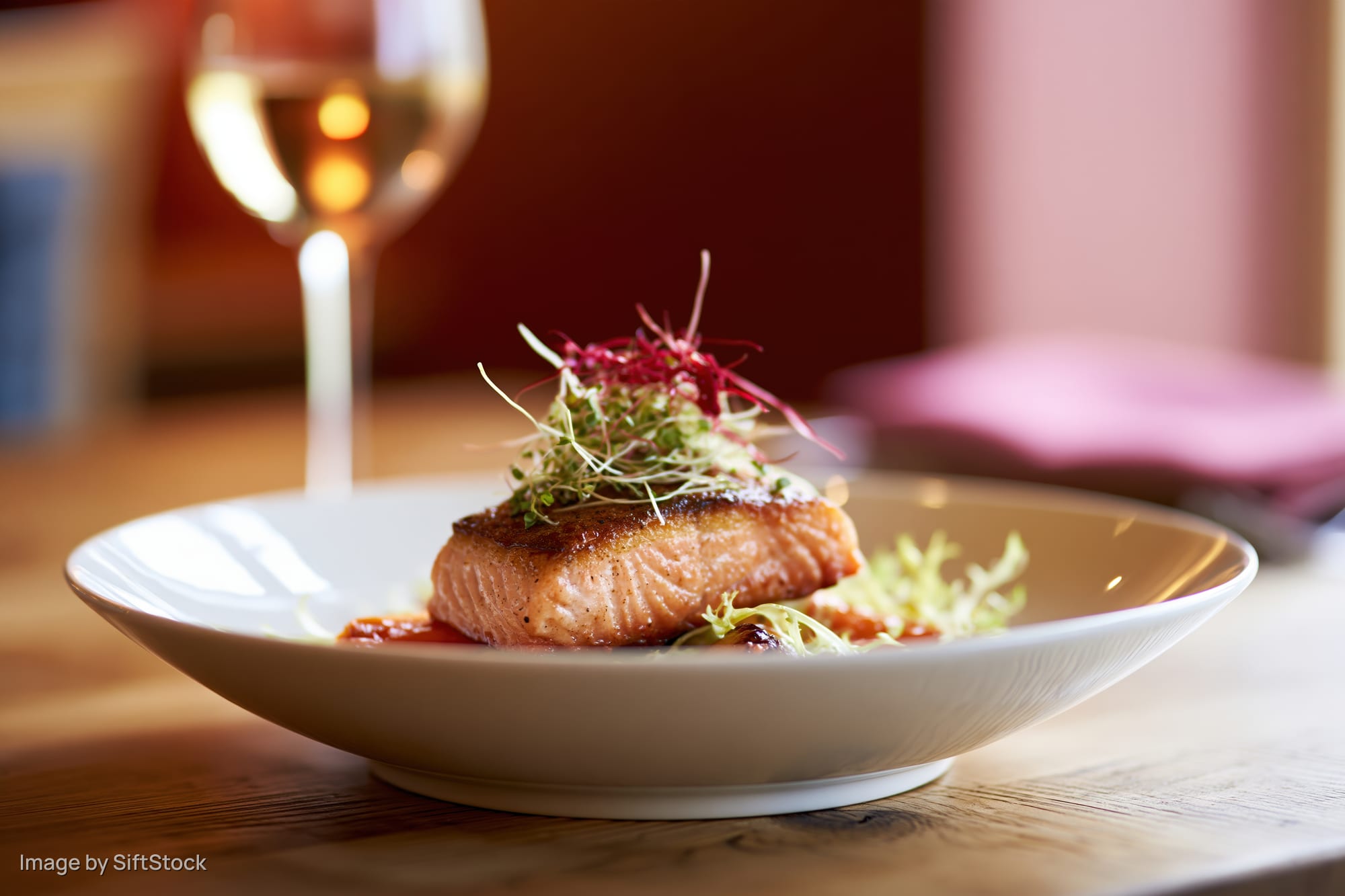 Seared salmon fillet topped with microgreens and red garnish on a white plate on a wooden table with a wine glass in the background