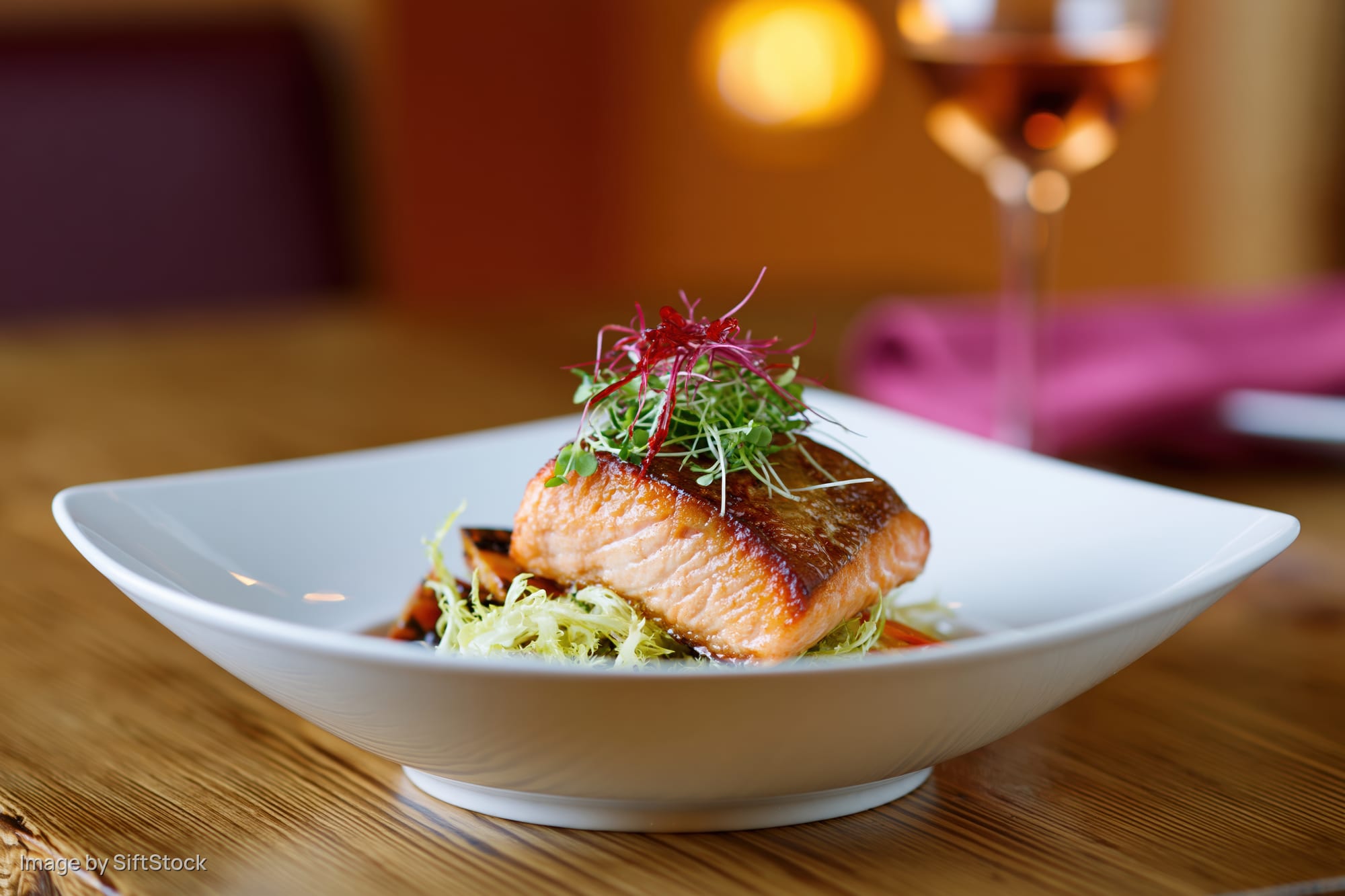 Seared salmon fillet topped with microgreens and red garnish on a white plate at a restaurant table
