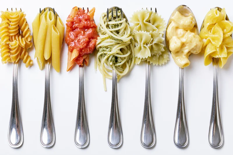Seven forks and spoons each holding different pasta types and sauces arranged in a row on white background