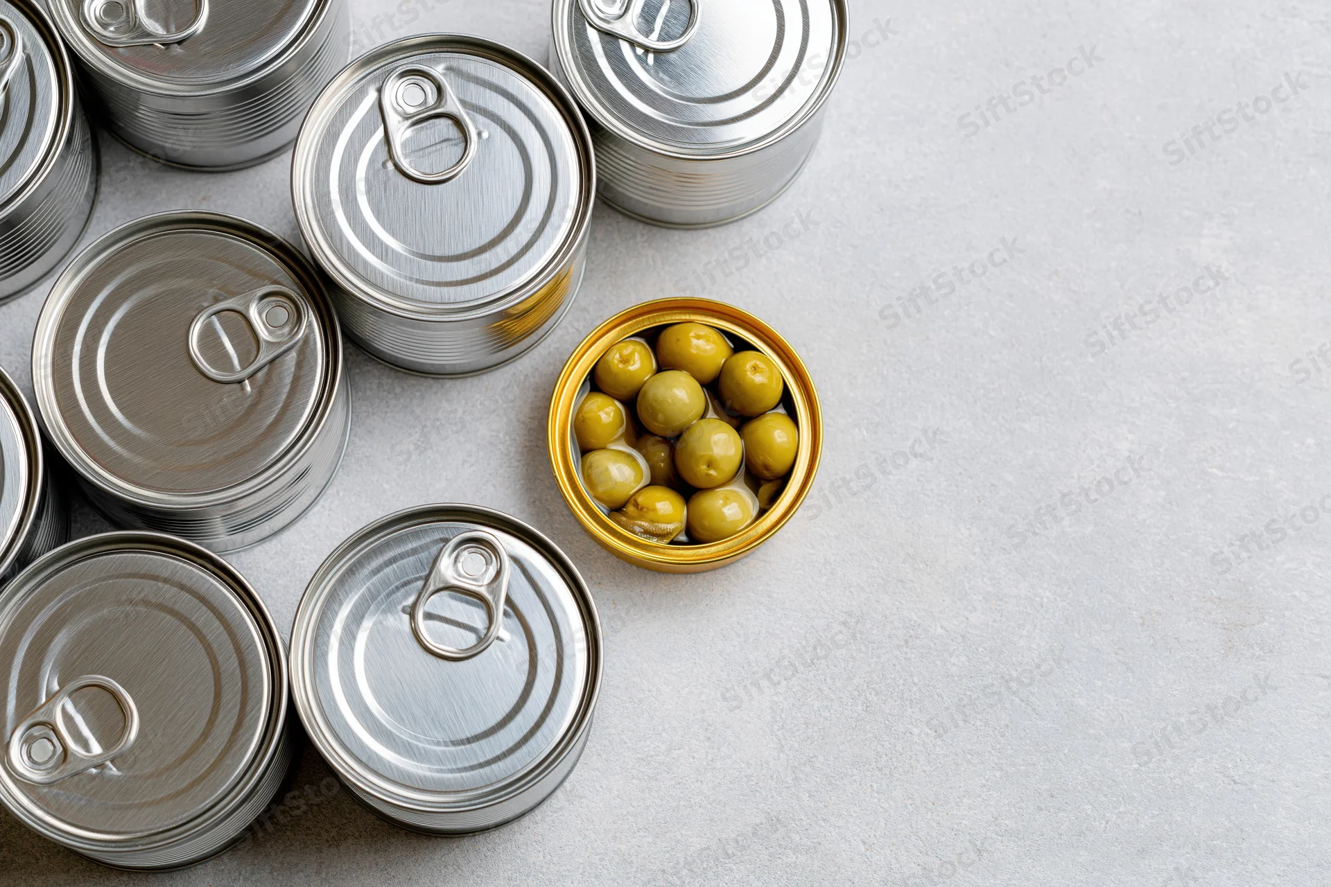Open tin can with green olives among closed silver cans on gray surface