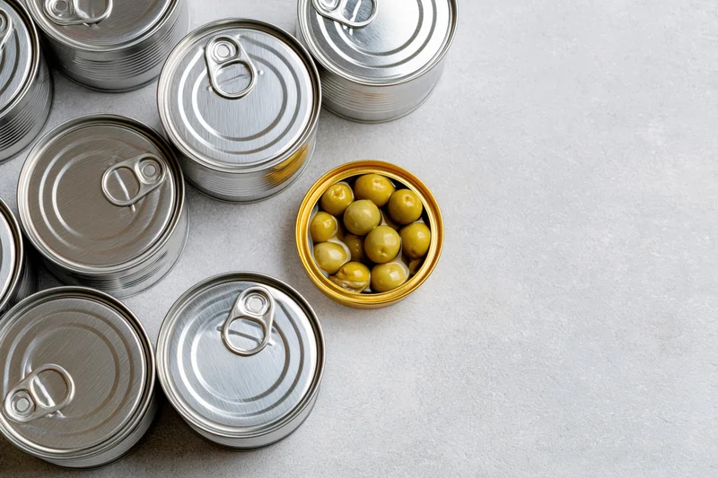 Open tin can with green olives among closed silver cans on gray surface