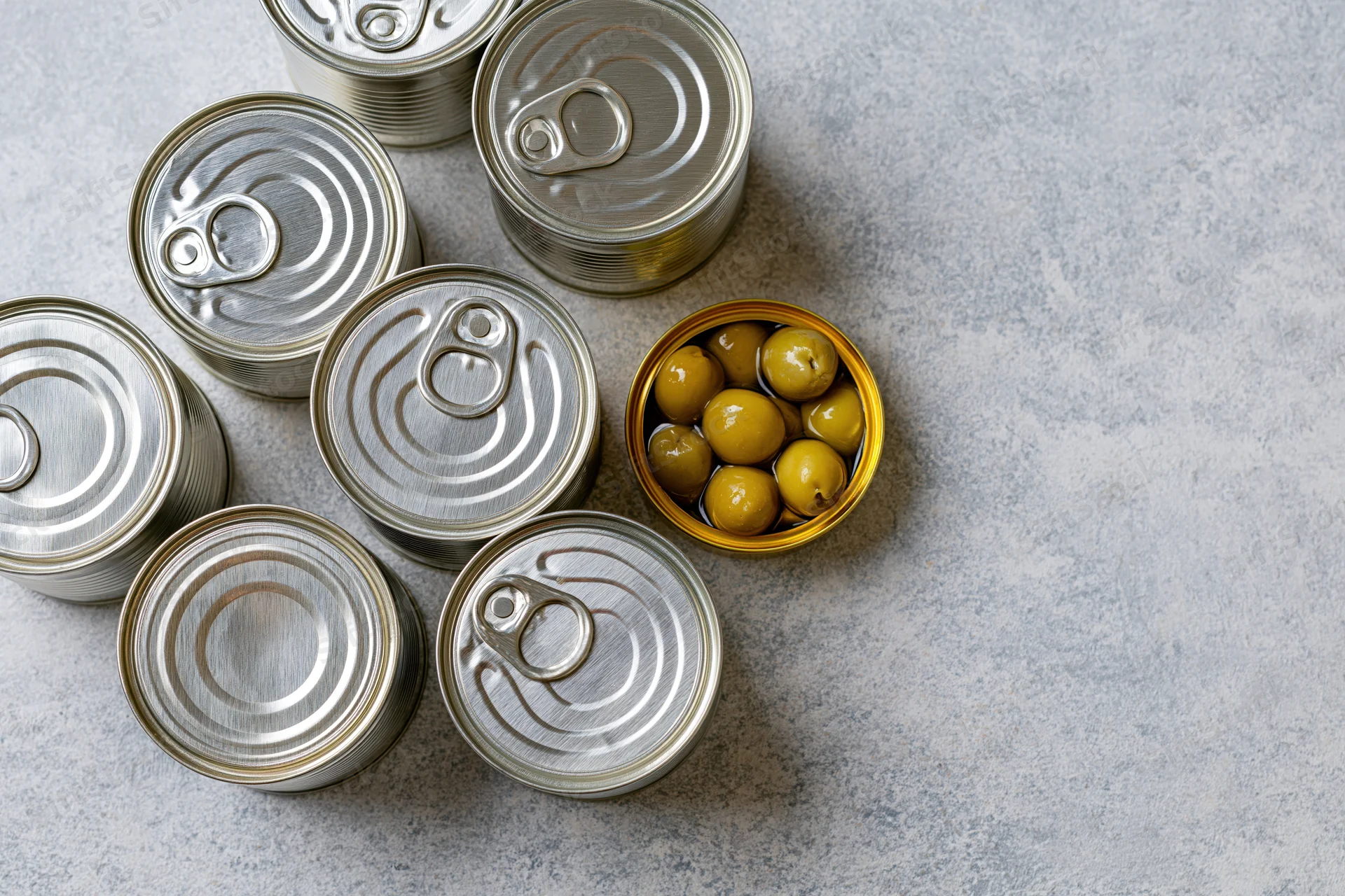 Several closed silver tin cans arranged on a flat surface with one open can revealing green olives inside