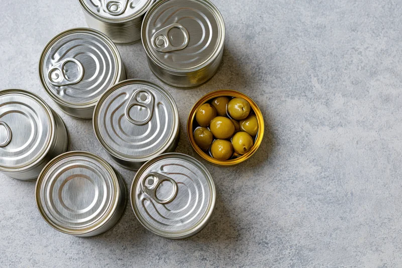 Several closed silver tin cans arranged on a flat surface with one open can revealing green olives inside
