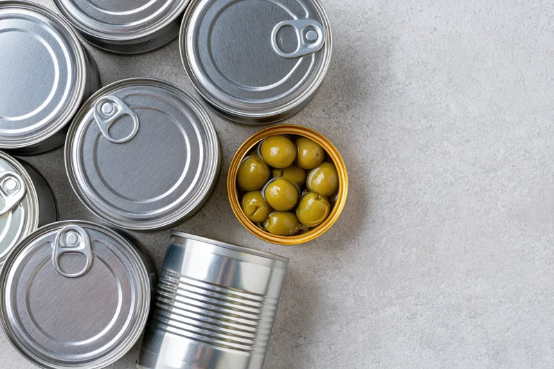 Several closed tin cans and one open can of green olives arranged on a gray countertop in a flat lay view