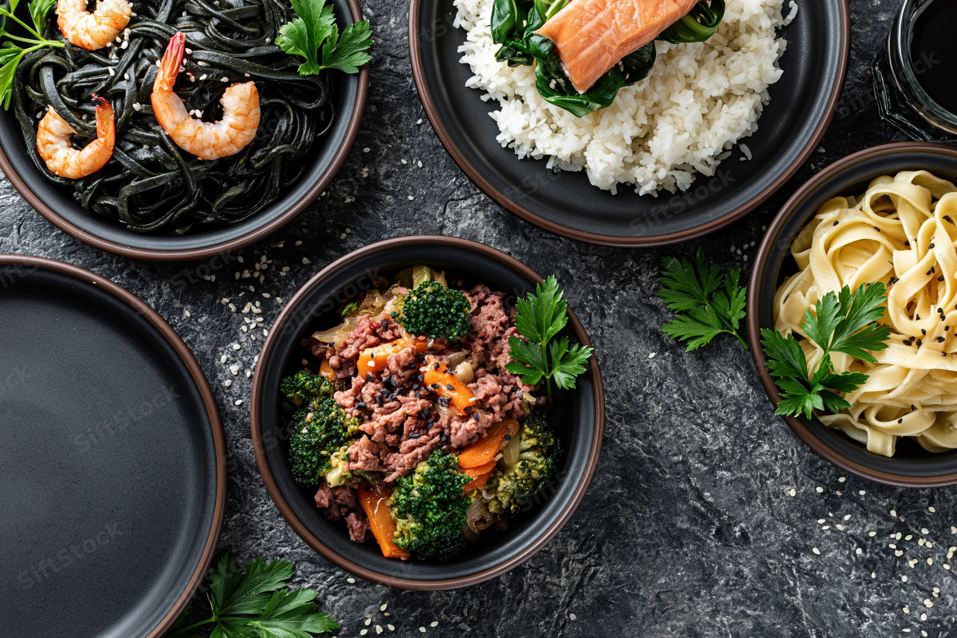 Several dark ceramic plates with different meals arranged on a black textured stone surface
