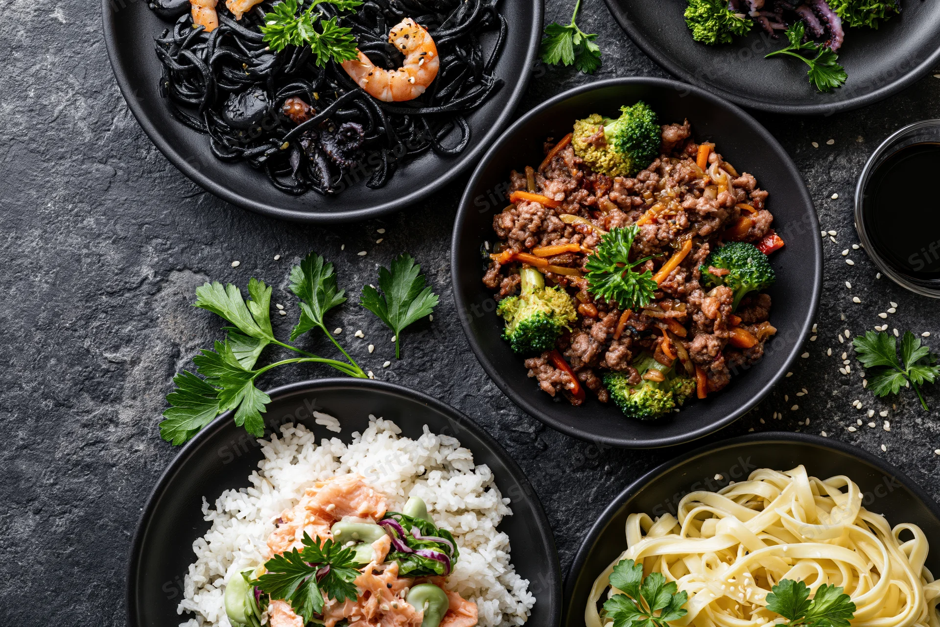 Several dark ceramic plates with different meals arranged on a black textured surface