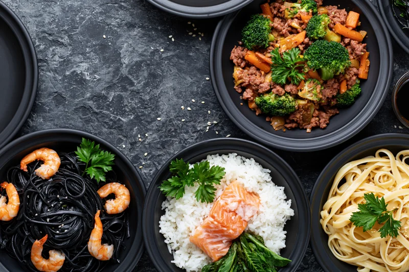 Several black ceramic bowls filled with different meals arranged on a dark textured surface