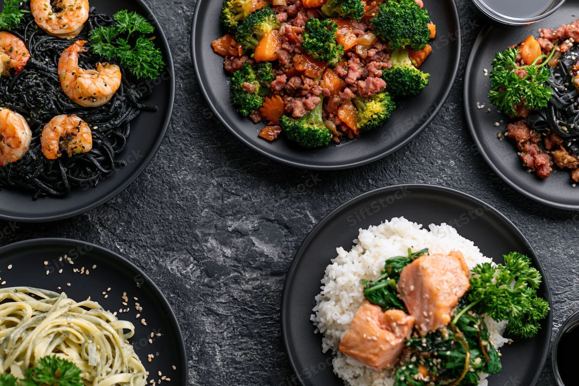 Several dark ceramic plates with different meals arranged on a black textured surface