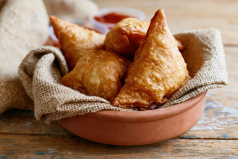 Golden-brown fried samosas piled in a terracotta bowl lined with burlap on a wooden table