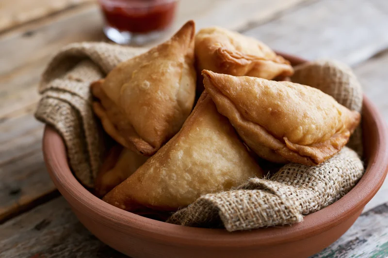 Golden-brown fried samosas piled in a terracotta bowl lined with burlap on a wooden table